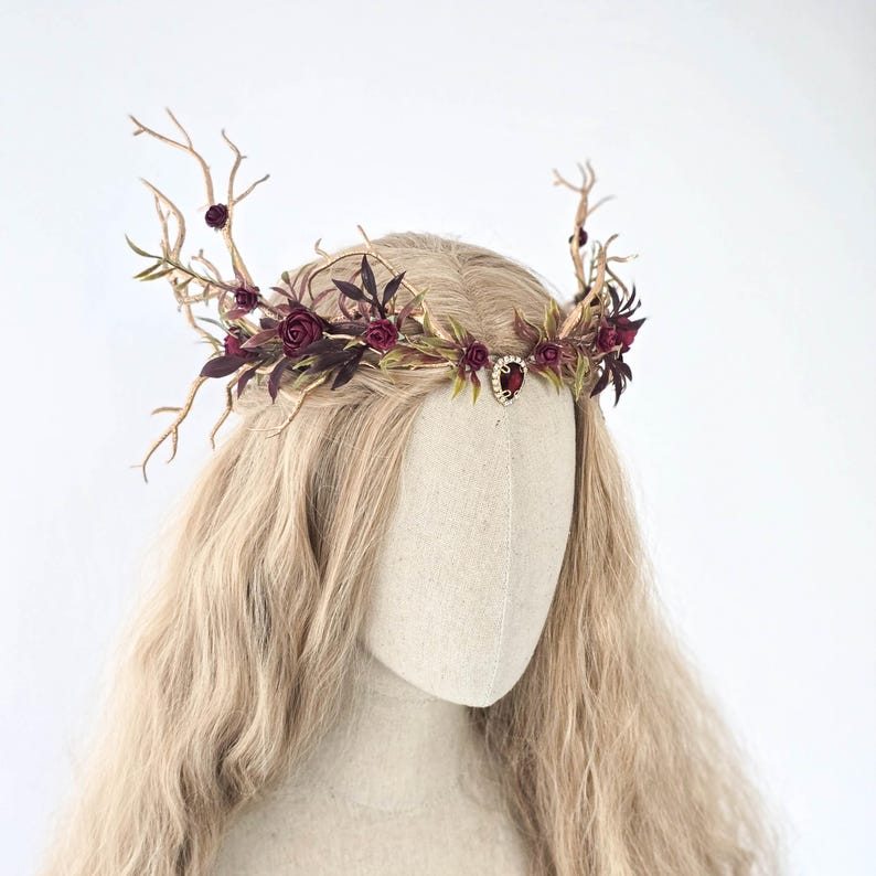 May include: A floral crown with burgundy roses, green leaves, and gold-toned branches. A small, ornate jewel is centered on the crown. The crown is displayed on a mannequin head with long, blonde hair, against a white background.