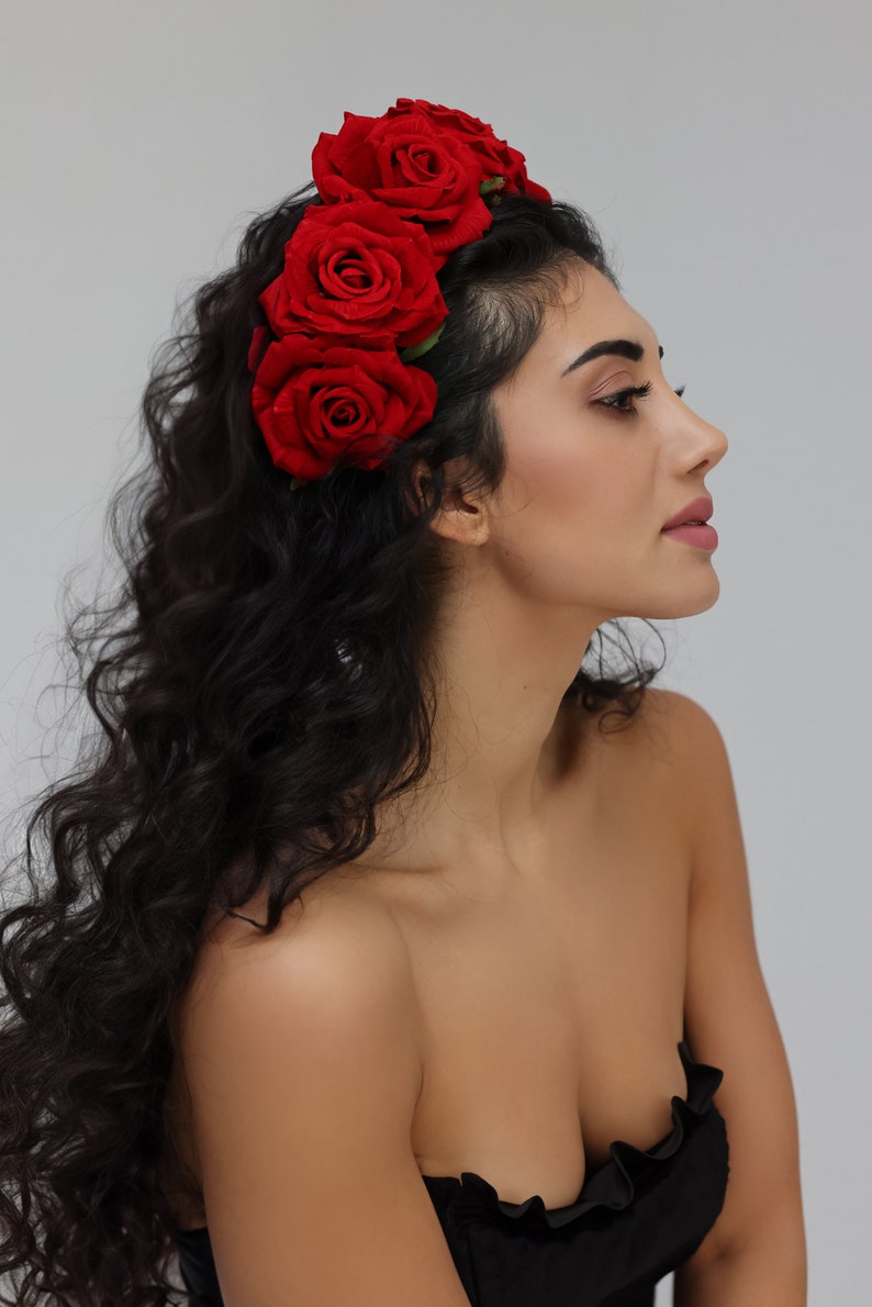 May include: A woman with long, curly black hair wearing a headband with red roses. She is wearing a black strapless top and has a natural makeup look.