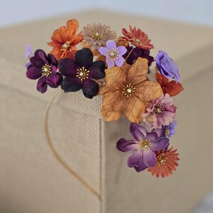 May include: A floral crown featuring an array of colorful fabric flowers. The flowers are in shades of orange, purple, brown, and lavender, with gold-colored accents in the center of each bloom. The crown is designed for special occasions.