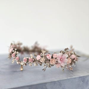 May include: A floral crown featuring pink and cream-colored fabric flowers and metallic leaves. The crown has a delicate design with small pearl accents and a dangling gold-colored charm. The flowers are arranged around the crown's base.