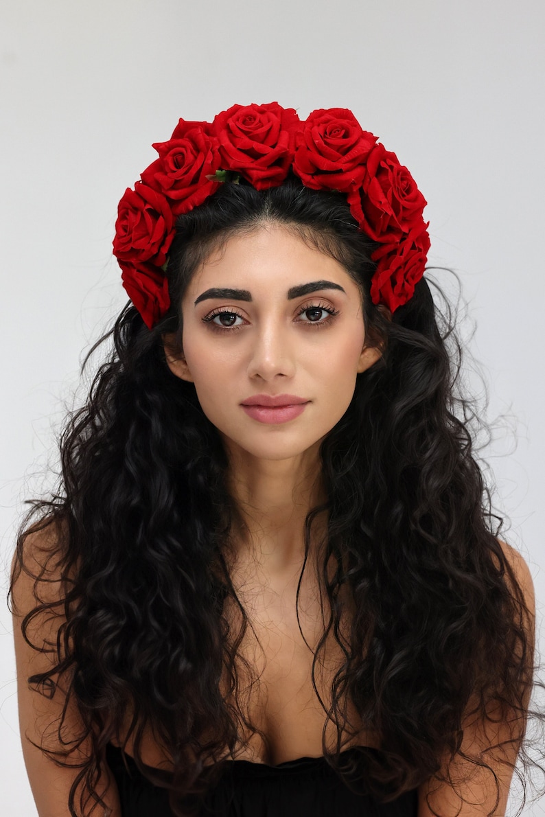 May include: A woman with long, curly black hair wearing a red rose flower crown.