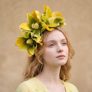 May include: A woman with a floral headpiece. The headpiece is made of large, layered flowers in yellow and green hues. The woman has long, wavy blonde hair and wears a light yellow dress. The background is a plain, neutral colour.
