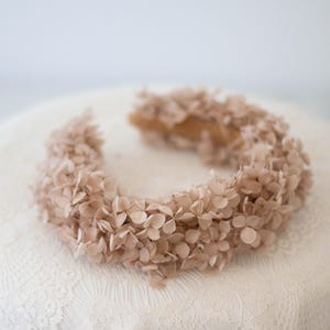 May include: A floral crown with light brown flowers. The crown is arranged in a circular shape, suitable for weddings or special events. The flowers are closely packed, giving a full and textured look. The crown rests on a white lace surface.