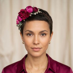 以下が含まれることがあります： 深紅とマゼンタ色の布製の花と金色の葉をあしらったフローラルヘッドバンド。花は細い濃い茶色のバンドにまとめられています。ヘッドバンドは、短い黒髪とバーガンディのシャツを着た人が着用しています。