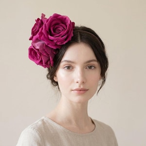 Op de afbeelding: Een persoon met donker haar draagt een haarband versierd met drie grote, levendige magenta rozen. De rozen zijn het middelpunt, met hun gelaagde bloemblaadjes en rijke kleur. De haarband is een dunne, bruine band.