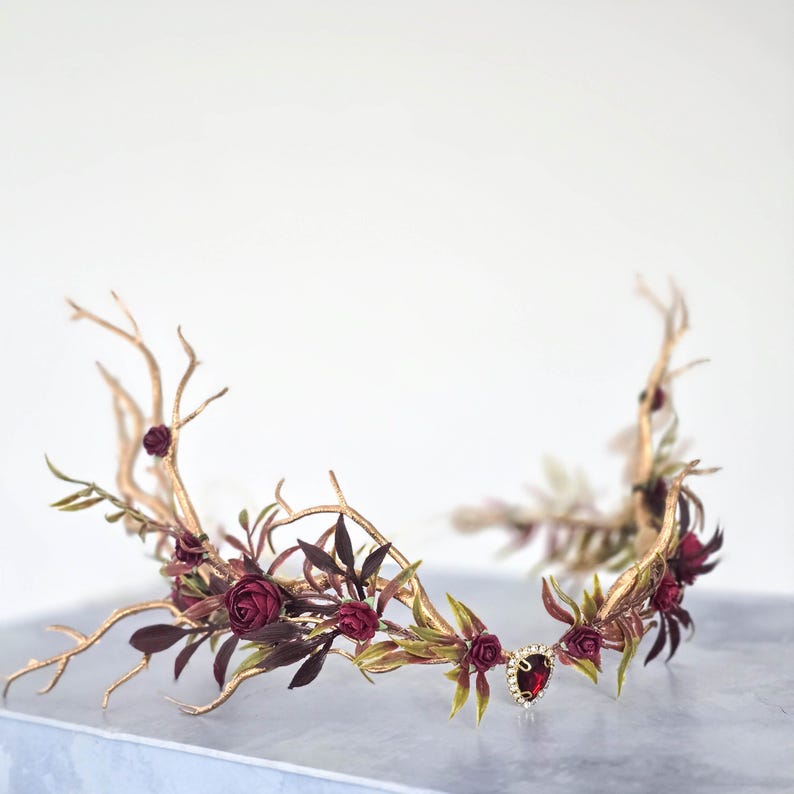 May include: A golden-toned floral crown with burgundy roses, green leaves, and dark burgundy foliage. The crown features a central teardrop-shaped jewel. The design incorporates branch-like elements, creating a natural, elegant aesthetic.