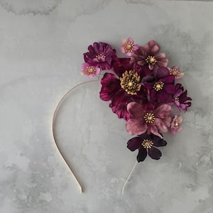 Puede incluir: Una diadema floral con un grupo de flores de tela de color púrpura oscuro, malva y rosa. Las flores están adornadas con detalles dorados y de cristal, sujetas a una fina banda metálica dorada. La disposición es asimétrica.