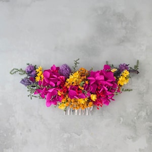 May include: A colourful floral hair comb with magenta, yellow, and purple flowers. The flowers are arranged on a silver comb, creating a beautiful accessory for special occasions.