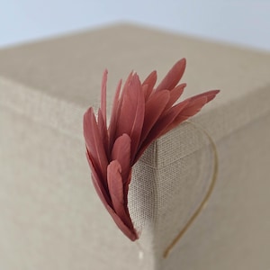 Può includere: Un copricapo con un gruppo di piume rosa antico. Le piume sono attaccate a una sottile fascia dorata. Il copricapo è posto su uno sfondo neutro e strutturato.