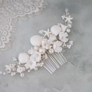 Op de afbeelding: Witte bruidskapselkam met een schelp- en bloemendesign. De kam is versierd met witte schelpen, parels en witte bloemen.