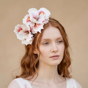 Op de afbeelding: Een bloemen haarband met drie grote, witte bloemen met roze en oranje accenten. De bloemen zijn bevestigd aan een dunne, goudkleurige metalen band. De haarband wordt gedragen door een persoon met lang, rood haar.
