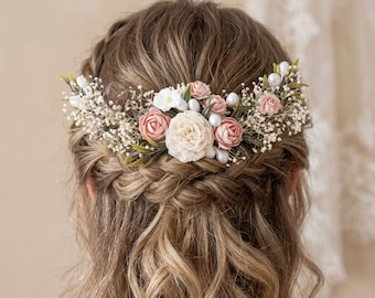 May include: A hair comb featuring a central cream-colored flower, surrounded by smaller dusty pink roses, white baby's breath, green leaves, and small white pearl beads. The floral arrangement is set on a comb base and worn in hair.