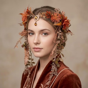 Può includere: Una donna indossa una corona floreale con accenti arancioni e marroni. La corona presenta un gioiello centrale ed elementi floreali a cascata. La donna indossa una veste di velluto color ruggine con dettagli ricamati.