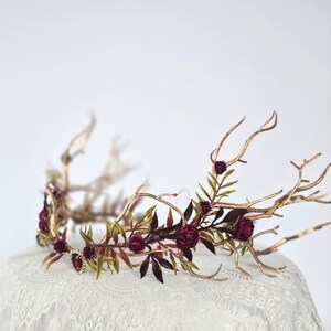 May include: A gold-toned antler crown with burgundy roses and green leaves. The floral crown features a central red jewel and is designed with a delicate, natural aesthetic. The crown is suitable for weddings, festivals, or themed events.