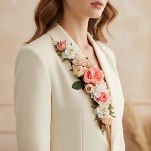 Può includere: Blazer color crema ornato da una composizione floreale di fiori rosa, bianchi e color pesca. I fiori scendono a cascata sul bavero, creando un accento decorativo. Il blazer ha un design classico.