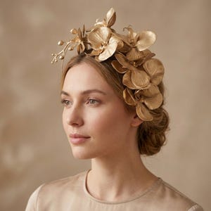Könnte beinhalten: Ein goldfarbener floraler Kopfschmuck bestehend aus einer Ansammlung geformter Orchideenblüten und zarten Stielen mit Knospen. Das metallische Accessoire ist asymmetrisch angeordnet, wobei mehrere geschichtete Blütenblätter eine texturierte Optik erzeugen.