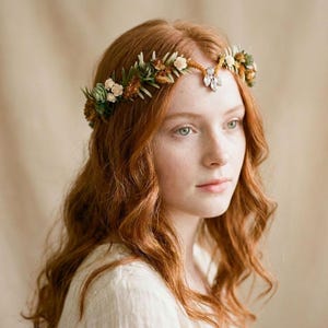 Könnte beinhalten: Ein floraler Haarkranz mit kleinen getrockneten Blumen und Sukkulenten in Grün-, Creme- und Brauntönen. Der Kranz wird auf dem Kopf getragen und hat eine Boho-Ästhetik. Ein kleiner Schmuckstein befindet sich in der Mitte der Stirn.
