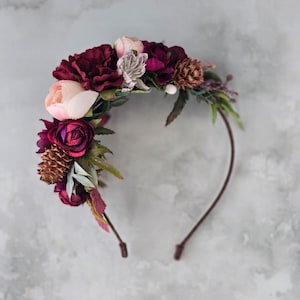 Op de afbeelding: Een dunne bruine haarband versierd met een arrangement van kunstbloemen en dennenappels. Het bloemstuk bevat diepbordeauxrode, zachtroze en witte bloesems, geaccentueerd met groene bladeren en kleine bruine dennenappels aan één kant.