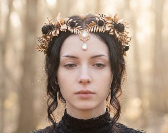 May include: A headpiece featuring gold-toned metallic leaves and small black roses arranged in a crown shape. A central gold-toned ornament sits at the forehead, adorned with a clear, iridescent teardrop-shaped crystal pendant hanging from it.