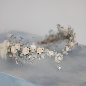 Op de afbeelding: Een delicate zilverkleurige bruidskroon met crèmekleurige rozen, kristallen accenten en een druppelvormige hanger. De bloemenhoofdband staat tegen een zachte blauwe achtergrond, ideaal voor bruiloften of speciale gelegenheden.