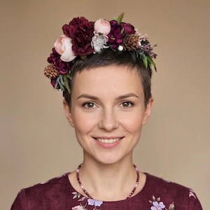 Könnte beinhalten: Eine Frau mit einem Blumenkranz mit weinroten, rosa und weißen Blumen und Tannenzapfen. Sie trägt ein weinrotes Kleid mit Blumenmuster und eine passende Perlenkette. Der Hintergrund ist einfarbig beige.