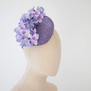 Op de afbeelding: Een paarse stoffen hoed met een ronde vorm en een tros lichtpaarse en blauwe bloemen erop.