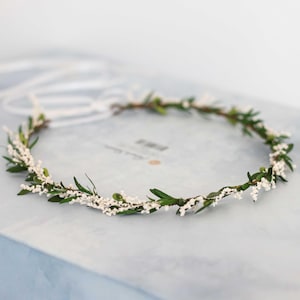 Puede incluir: Una corona de flores blancas con hojas verdes. La corona está hecha de pequeñas flores blancas y hojas verdes.