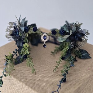 Pode incluir: Uma coroa de flores artesanal com rosas azuis escuras, folhagem verde e um pendente central de joia. A coroa &eacute; adornada com v&aacute;rias folhas e bagas, criando uma est&eacute;tica elegante e natural. Perfeita para casamentos ou ocasi&otilde;es especiais.