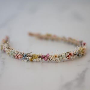 Puede incluir: Una delicada diadema floral con una variedad de pequeñas flores coloridas en tonos rosa, amarillo, azul y blanco. Las flores están dispuestas alrededor de una fina banda beige, creando un accesorio natural y elegante.