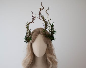 May include: A decorative headpiece featuring a crown of brown, twisting branches adorned with green foliage. The crown sits atop a mannequin head with long, wavy blonde hair, set against a plain white background.