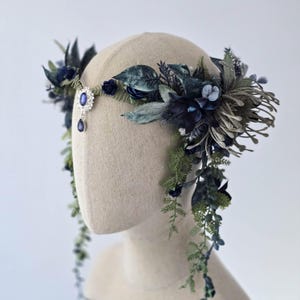Pode incluir: Uma grinalda floral com flores e folhagens artificiais azuis escuros e verdes. Uma joia central com um detalhe de l&aacute;grima pendurada acentua o design. A grinalda &eacute; exibida em uma cabe&ccedil;a de manequim, mostrando seus detalhes.