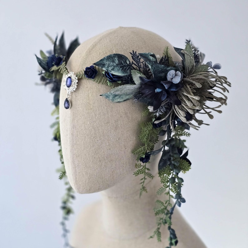 Pode incluir: Uma grinalda floral com flores e folhagens artificiais azuis escuros e verdes. Uma joia central com um detalhe de l&aacute;grima pendurada acentua o design. A grinalda &eacute; exibida em uma cabe&ccedil;a de manequim, mostrando seus detalhes.