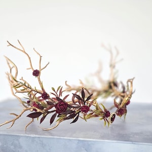 May include: A gold-colored crown with a twig-like design, adorned with burgundy roses and green and burgundy leaves. The crown is set against a white background, creating a striking contrast. The floral elements add a touch of elegance.