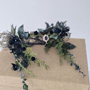 Pode incluir: Uma coroa floral azul escuro com um pendente de joia central. A coroa apresenta rosas azul escuro, folhagem verde e detalhes prateados. A coroa &eacute; exibida em uma superf&iacute;cie marrom.