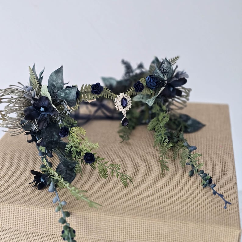 Pode incluir: Uma coroa floral azul escuro com um pendente de joia central. A coroa apresenta rosas azul escuro, folhagem verde e detalhes prateados. A coroa &eacute; exibida em uma superf&iacute;cie marrom.