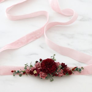 May include: A pink velvet ribbon with a burgundy floral embellishment. The embellishment features a variety of burgundy and pink flowers, greenery, and berries.