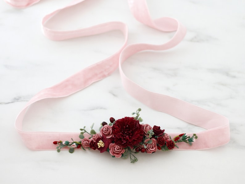 May include: A pink velvet ribbon with a burgundy floral embellishment. The embellishment features a variety of burgundy and pink flowers, greenery, and berries.