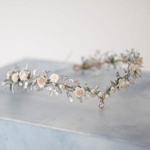 May include: A floral crown with cream roses and silver leaves. The headpiece is decorated with small white flowers and a crystal charm. Ideal for weddings or special events, it measures approximately 20 cm in diameter.