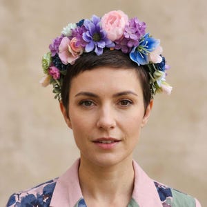Könnte beinhalten: Eine Frau mit kurzen, dunklen Haaren trägt einen Blumenkranz mit rosa, lila und blauen Blüten. Sie trägt eine Jacke mit rosa Kragen und blauem Blumenmuster.