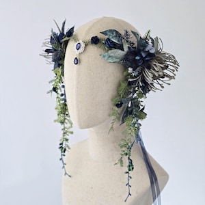 Pode incluir: Uma grinalda floral com flores e folhagens artificiais azuis escuros e verdes. Apresenta uma joia central e vegeta&ccedil;&atilde;o pendurada, com uma fita azul. O design &eacute; exibido em uma cabe&ccedil;a de manequim.