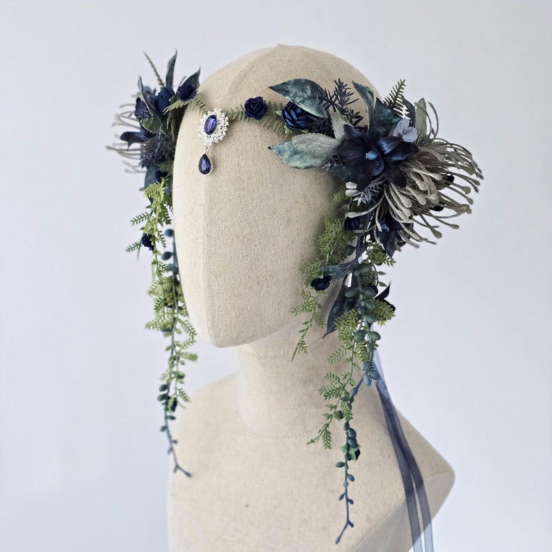 Pode incluir: Uma grinalda floral com flores e folhagens artificiais azuis escuros e verdes. Apresenta uma joia central e vegeta&ccedil;&atilde;o pendurada, com uma fita azul. O design &eacute; exibido em uma cabe&ccedil;a de manequim.