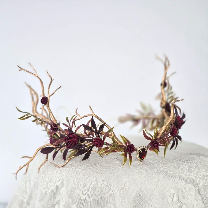 May include: A handcrafted crown featuring gold-colored branches, burgundy roses, and green leaves. A sparkling red gem is set at the center. The floral crown is designed for special occasions.