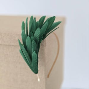 Pode incluir: Uma tiara de penas verdes com uma faixa dourada. As penas são de um tom verde profundo e estão dispostas em forma de leque. A tiara está sobre uma superfície bege.