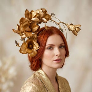May include: A gold-coloured headband featuring a cluster of metallic gold magnolia flowers and thin branches. The floral arrangement is positioned on one side of the headband, showcasing detailed petals and small buds with a textured, shimmering finish.