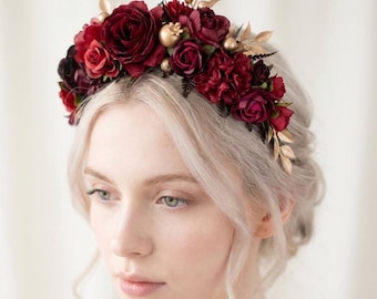 May include: A floral crown featuring deep red roses, smaller burgundy flowers, and gold accents. The crown is adorned with gold-colored leaves and small golden spheres. The flowers are arranged on a dark base, creating a striking contrast.