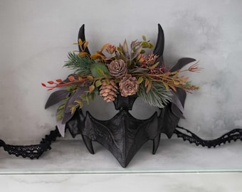May include: A black masquerade mask with horns and a floral arrangement of pine cones, leaves, and berries.