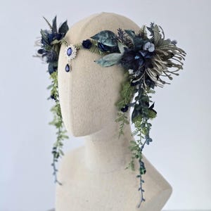Pode incluir: Uma grinalda floral com folhas e flores azuis escuras e verdes. Um pingente prateado com uma gema azul pende do centro. A grinalda &eacute; exibida em uma cabe&ccedil;a de manequim.