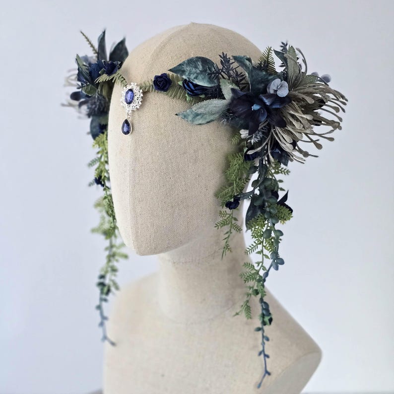 Pode incluir: Uma grinalda floral com folhas e flores azuis escuras e verdes. Um pingente prateado com uma gema azul pende do centro. A grinalda &eacute; exibida em uma cabe&ccedil;a de manequim.
