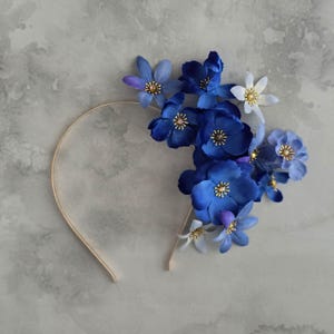 Könnte beinhalten: Ein floraler Haarreif mit einem goldfarbenen Metallband, verziert mit einem Cluster aus blauen Stoffblumen. Die Blumen variieren in Blautönen, mit goldenen Zentren, und einige weiße Blumen sind in der Anordnung verstreut.