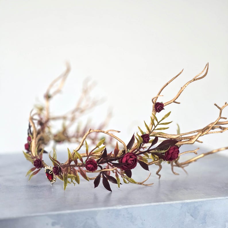 May include: A gold-toned crown with a branch-like design, adorned with burgundy roses and green leaves. A small, red jewel is set at the center. The crown is a decorative headpiece.